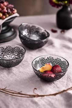 three glass bowls with fruit in them sitting on a white table cloth next to flowers