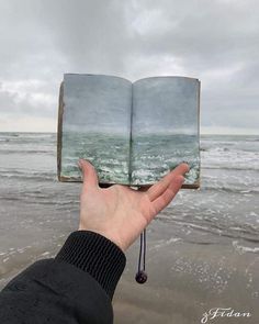 a person holding an open book in front of the ocean on a cloudy day with waves