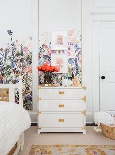 a white dresser sitting next to a bed in a room with flowers on the wall