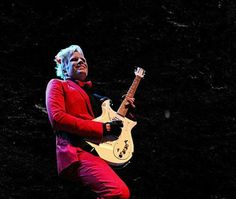 the man is playing an electric guitar in his red suit and black shirt with white hair