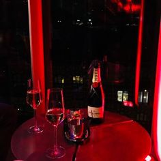 two wine glasses and a bottle on a table in front of a window with red lights