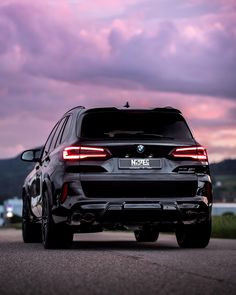 the rear end of a black bmw suv parked in front of a purple sky with clouds