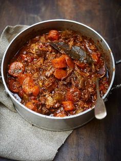 a stew with carrots and meat in a pot on a wooden table next to a spoon