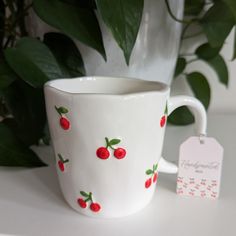 a white cup with cherries on it next to a potted plant and tag