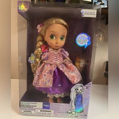 the doll is in its box and it has a purple dress with flowers on it