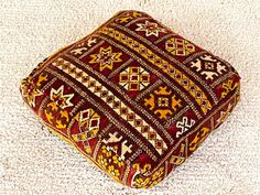 a small square cushion on the floor with white carpet in the background and an orange, yellow, red, and brown pattern