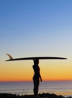 a woman holding a surfboard over her head while standing next to the ocean at sunset