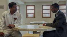 two men sitting at a table talking to each other in a room with tables and chairs