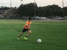 a young man kicking a soccer ball on top of a lush green field with buildings in the background