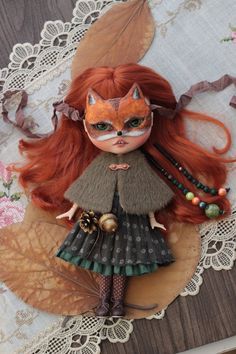 a doll with long red hair wearing a green dress and orange cat mask on top of a lace doily