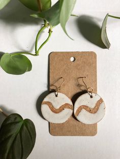 two white and brown earrings sitting on top of a piece of paper next to a plant