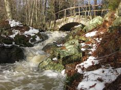 a bridge over a small stream in the woods with snow on the ground and rocks