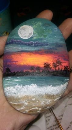 a hand holding a painted rock with a sunset and moon in the sky on it