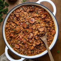 a pot filled with beans and sausage on top of a wooden table next to a spoon