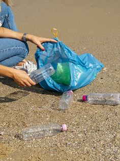 a woman sitting on the beach with plastic bottles in her hand and an empty bag next to it