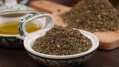 two bowls filled with different types of herbs next to a wooden spoon full of oil