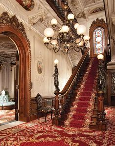 an ornate staircase with red carpet and chandelier in the center, leading up to another room