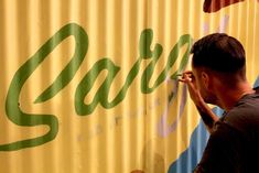 a man is painting the side of a sign on a yellow and blue wall with green lettering