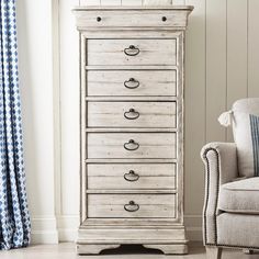 an old white chest of drawers next to a chair and window with blue drapes