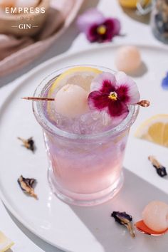 a small glass filled with liquid and flowers on top of a white plate next to lemon slices