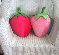 two stuffed strawberries sitting on a white wicker chair, one is pink and the other is green