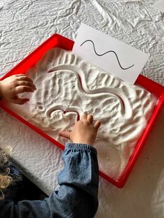 a child is playing with sand in a red tray on top of a white bed