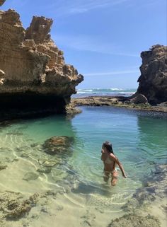 a woman wading in clear water near rocks and the ocean on a sunny day