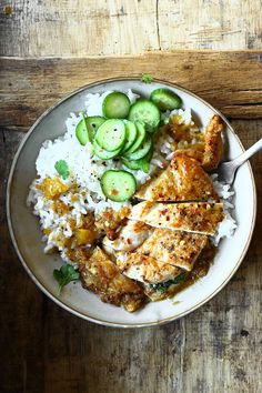 a white plate topped with chicken and rice next to cucumbers on top of a wooden table