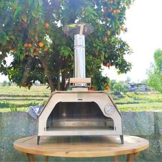 an outdoor pizza oven sitting on top of a wooden table next to a fruit tree