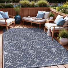 an outdoor area rug with blue and white pillows on top of wooden flooring, surrounded by potted plants