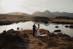 a bride and groom standing on the edge of a mountain overlooking a lake with mountains in the background