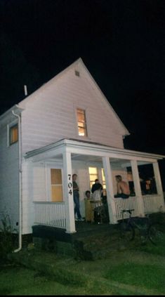 people are standing on the porch of a white house at night with their hands in their pockets