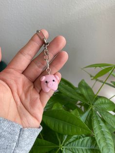 a hand holding a pink pig keychain in front of a potted plant