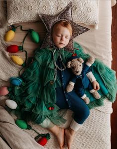 a baby sleeping on top of a bed next to a teddy bear wearing a green dress