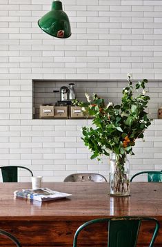 a wooden table topped with green chairs and a vase filled with flowers next to a brick wall