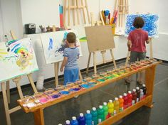 two young children are painting on canvass in an art room with easels and water bottles