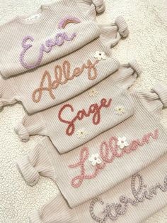 four sweaters with embroidered letters on them sitting on a bed together, all in different colors