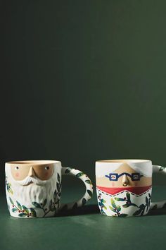 two coffee mugs with faces painted on them sitting next to each other in front of a green background