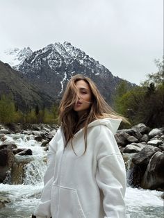 a woman standing in front of a mountain stream wearing a white hoodie and black pants