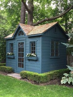 a small blue shed with windows and a wooden roof in the middle of a yard