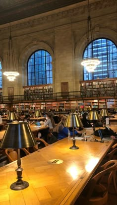 people sitting at tables in a large library with many bookshelves and lamps on them