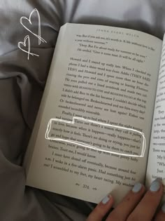 a person is reading a book on a bed with grey sheets and white nail polish