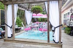 an outdoor dining area with white curtains and pink cushions on the table, surrounded by greenery