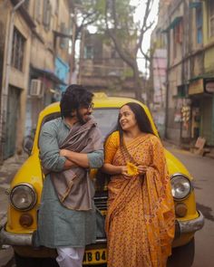 two people standing in front of a yellow car on a street with buildings behind them