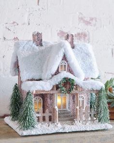 a small house with snow on the roof and trees around it, sitting on a table next to a potted plant