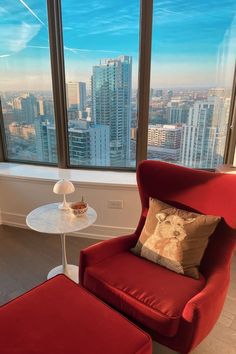 a red chair and ottoman in front of a large window with cityscape outside