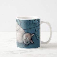 a white cat sleeping on top of a blue surface coffee mug with an image of a siamese cat