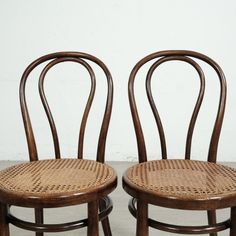 two wooden chairs sitting side by side in front of a white wall and one has a wicker seat