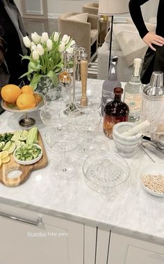 a woman standing next to a kitchen counter filled with food and wine glasses on top of it