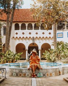 a woman in an orange dress is sitting on a bench near a pool and palm trees